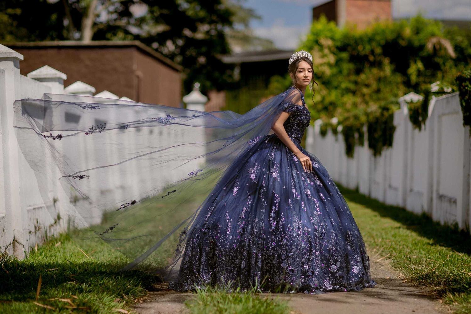 A sparkly navy blue ball gown shimmers in sunlight along a white picket fence on a grassy pathway.