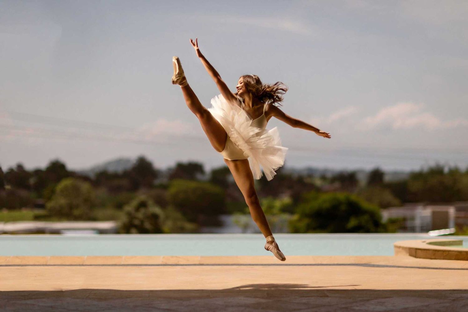 A ballet dancer in a white dress performs a graceful leap outdoors against a natural landscape backdrop.