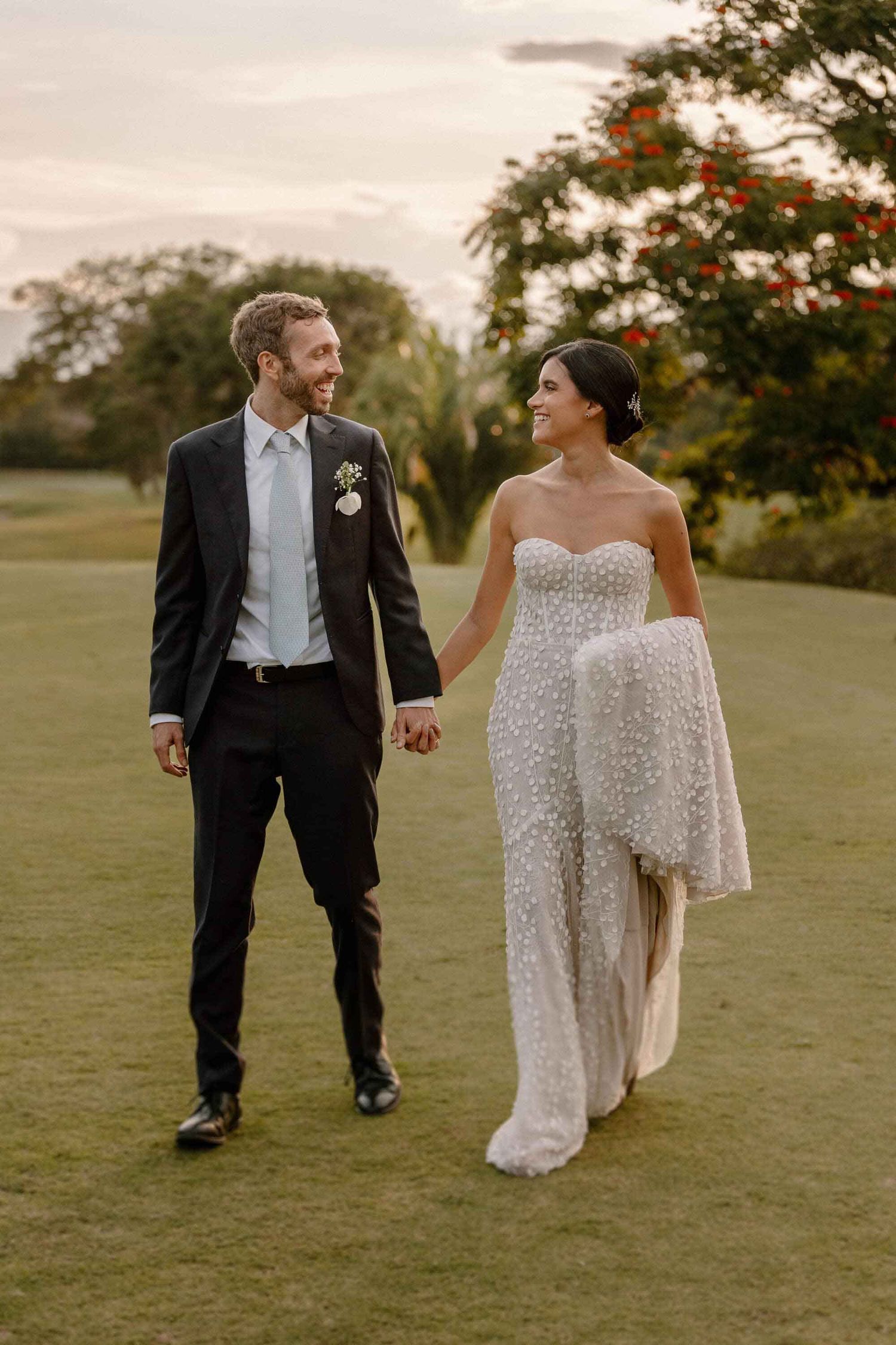 A newlywed couple walking hand in hand on a lawn during golden hour, with flowering trees in the background.