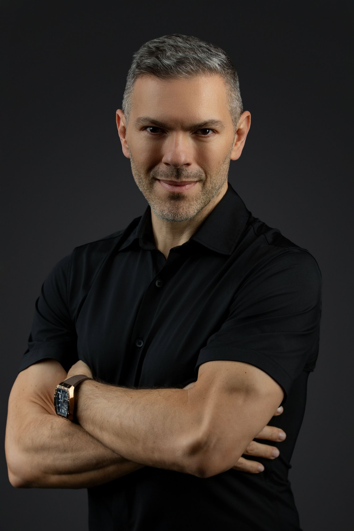 Professional studio portrait of a person wearing a black button-up shirt with arms crossed against dark background.