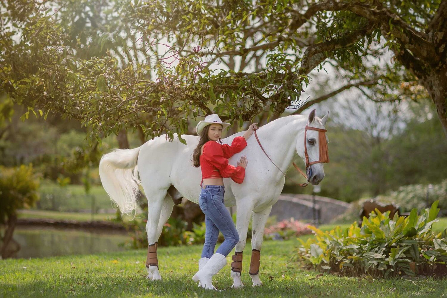 A person in a red sweater and jeans stands beside a majestic white horse in a lush garden setting.