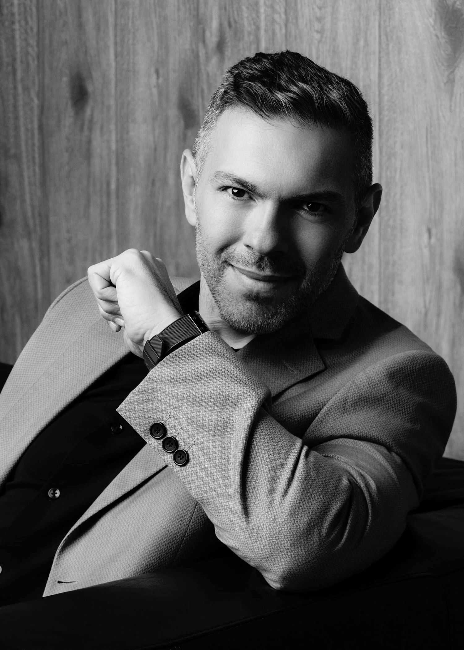 Black and white portrait of someone in a blazer smiling at the camera against a wooden wall background.