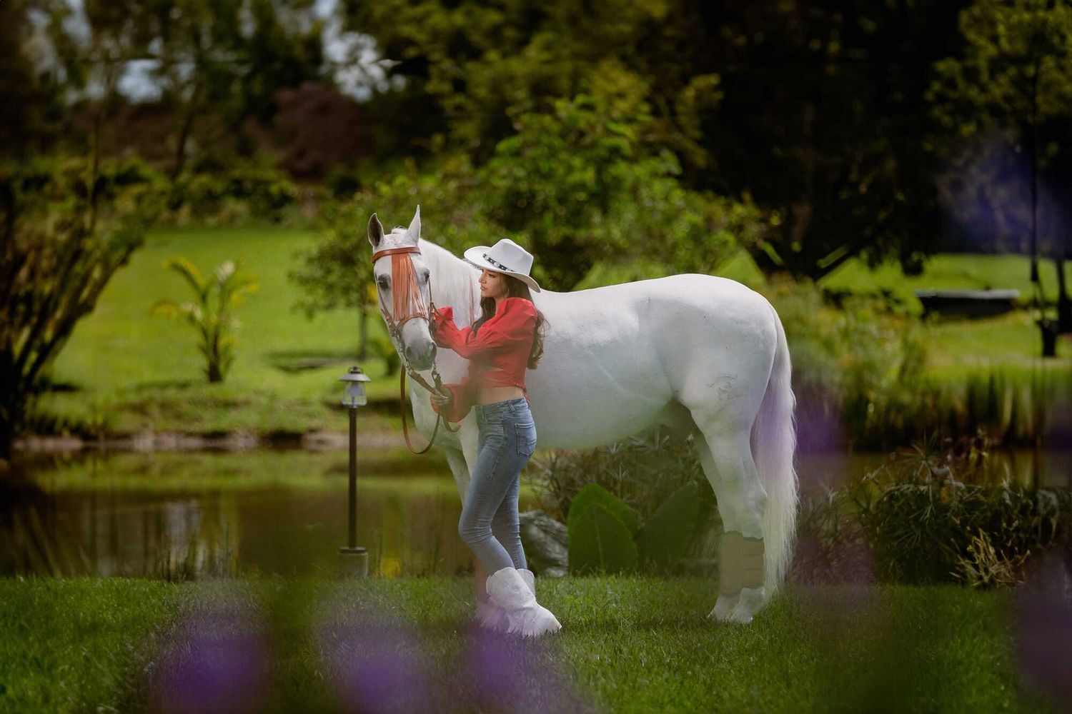 A person in red hugs and comforts a white horse in a tranquil garden setting beside a pond.