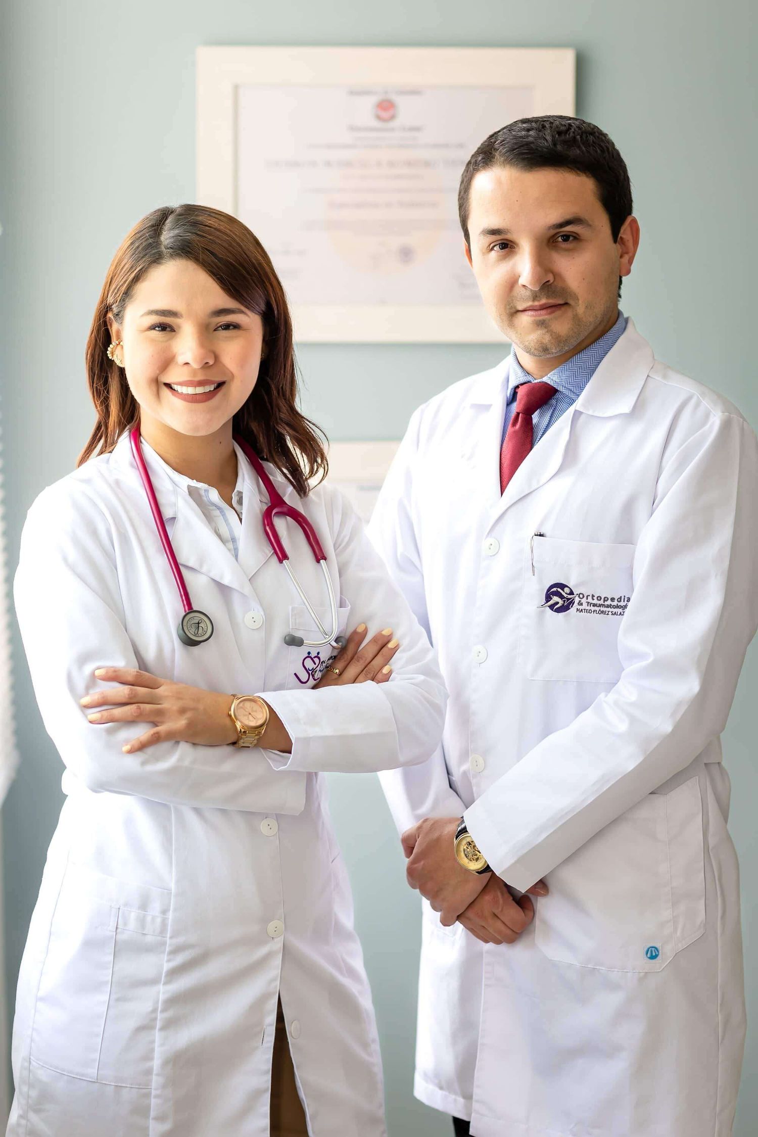 Two healthcare professionals in white lab coats and stethoscopes stand confidently in a medical office setting.
