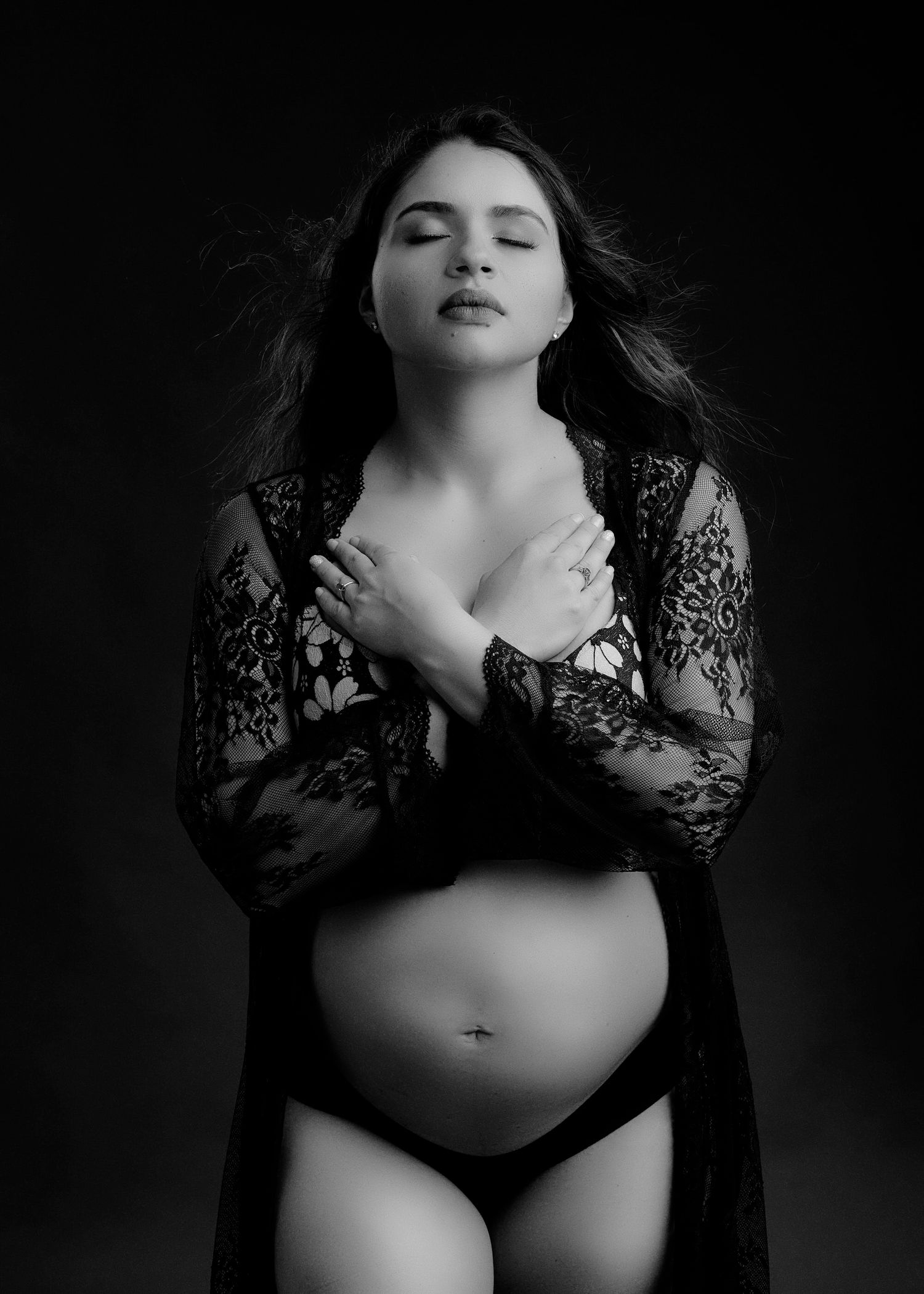 Black and white maternity photo of figure in lace robe and dark lingerie with windswept hair against dark background.