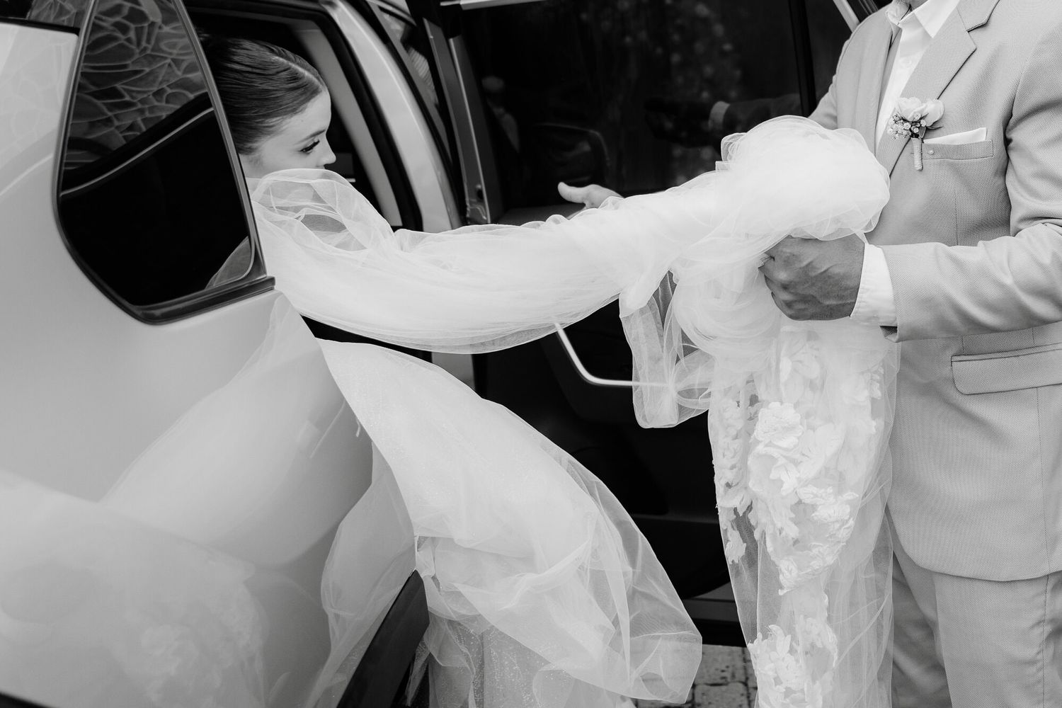 A close-up black and white photo shows someone helping lift a flowing white wedding dress into a car.