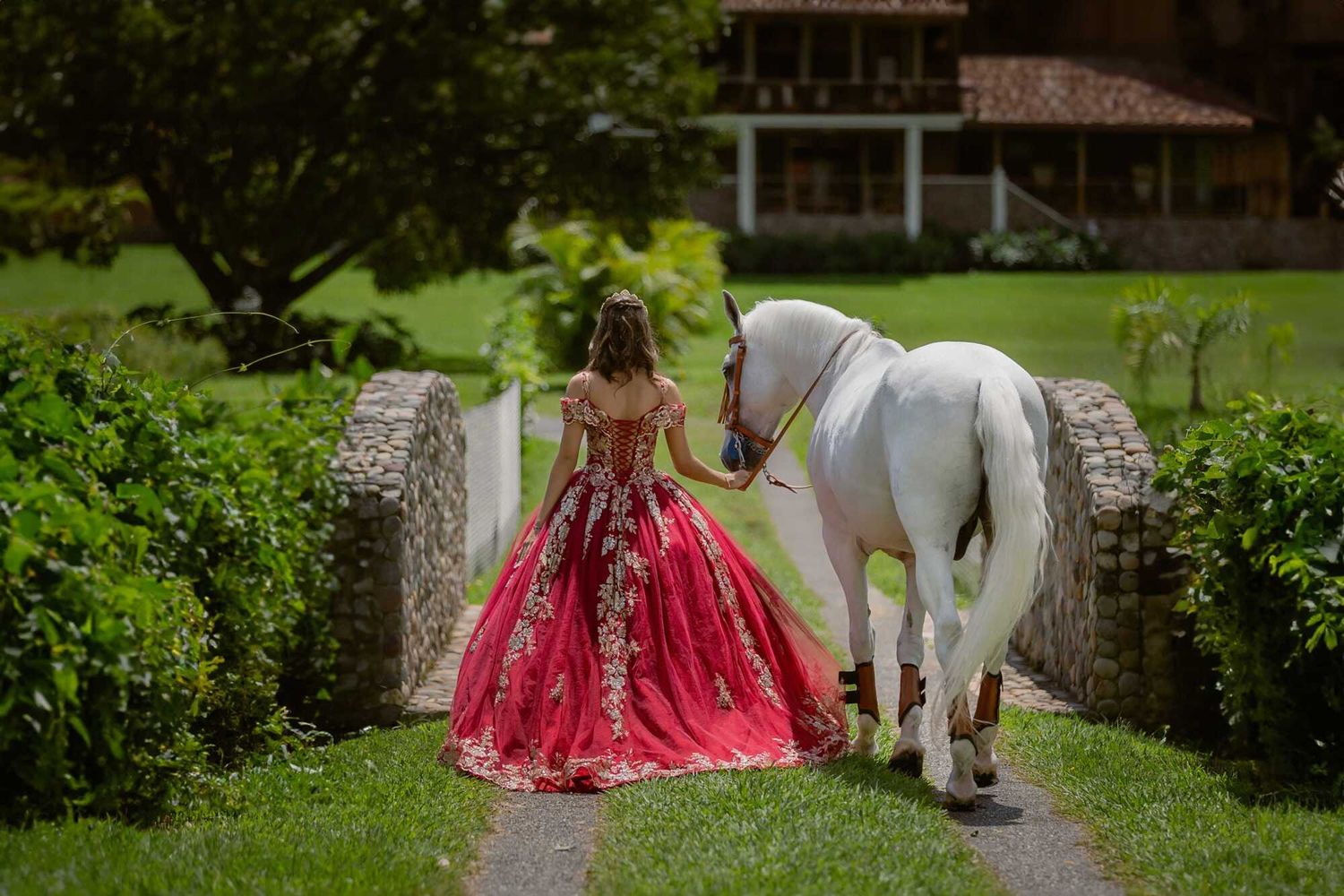A person in a vibrant pink formal gown stands on a stone bridge with a white horse in a lush garden setting.