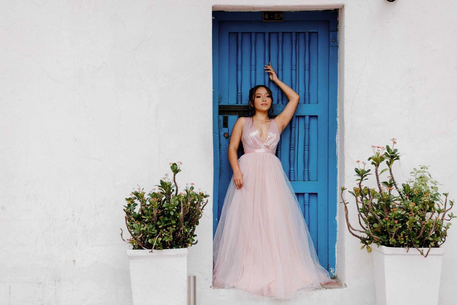 A flowing pink tulle gown stands out against a bright blue door and white stucco wall with potted plants.