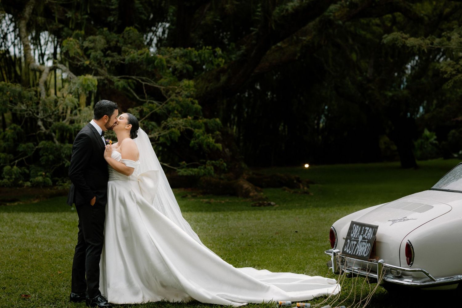 A romantic wedding photo captured outdoors with a classic vintage car and lush garden backdrop.