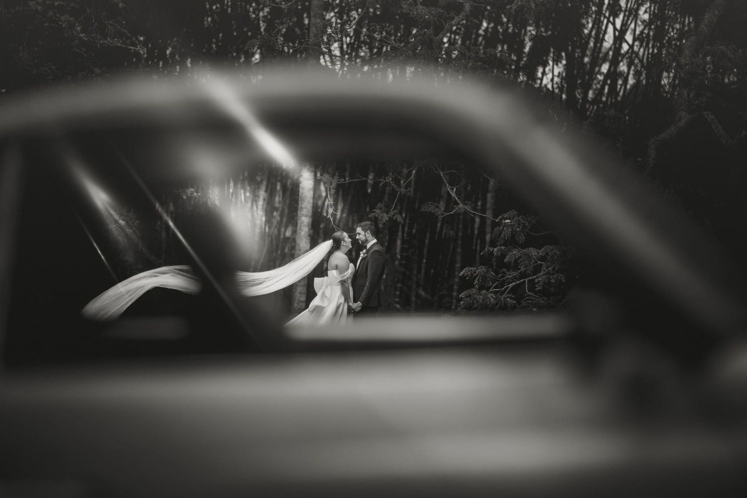 Foto de boda en Medellín: Novios besándose en el bosque con el velo al viento, captados a través de la ventana de un auto antiguo.