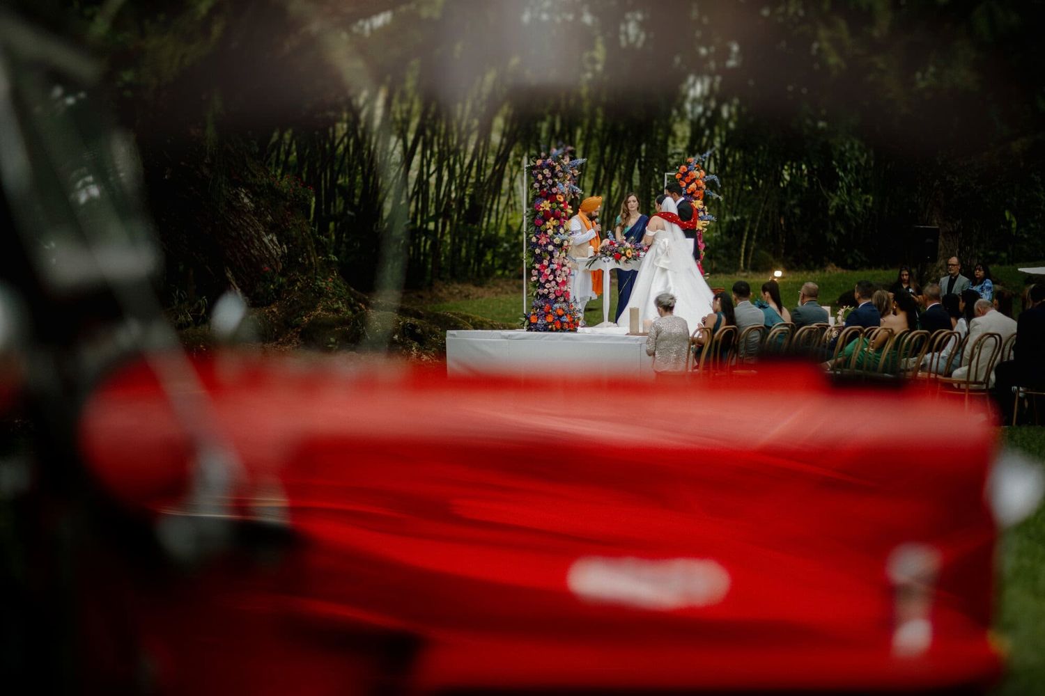 Outdoor wedding ceremony with red chairs in foreground, creating a unique perspective through blurred frame.
