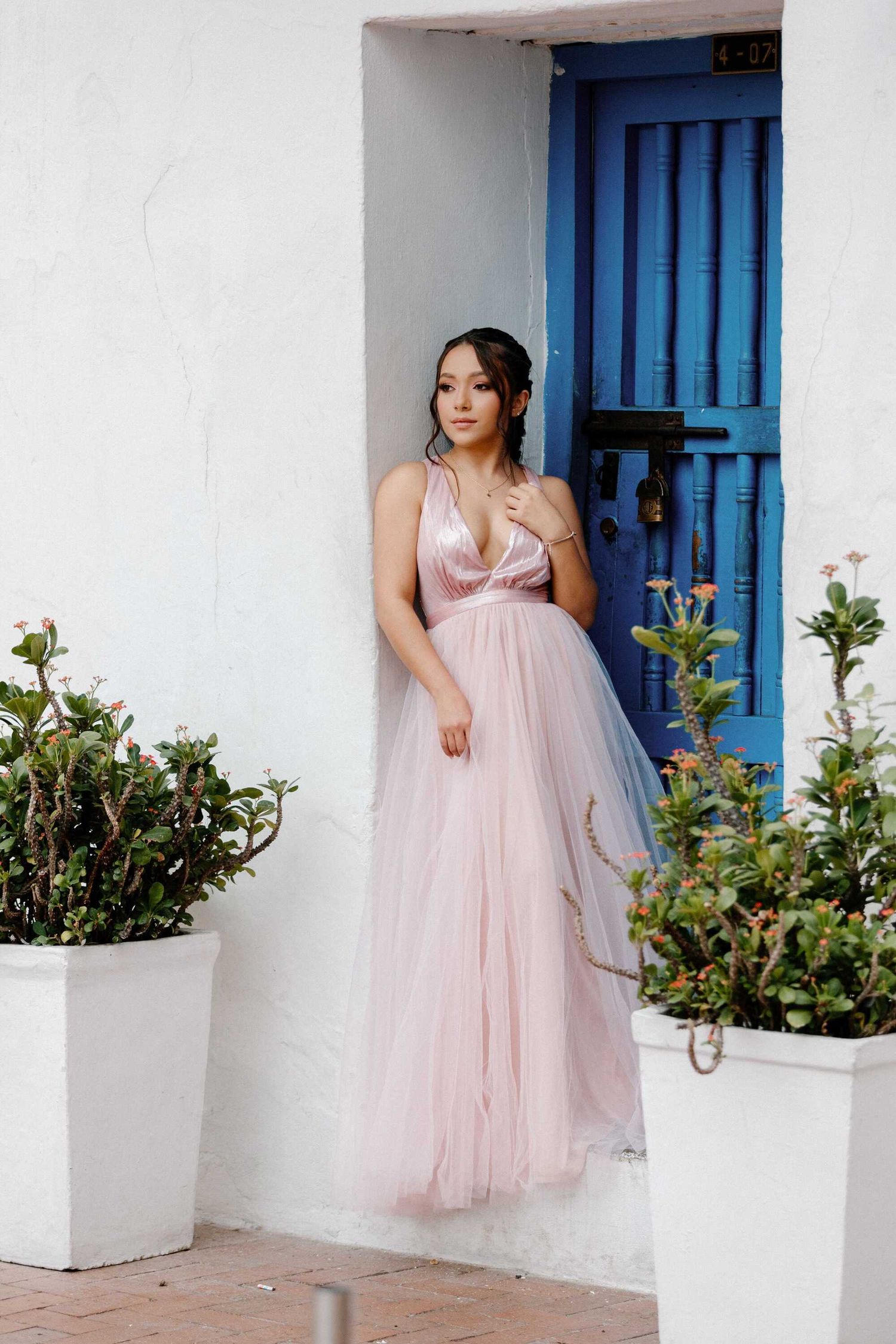 Woman in a flowy pink tulle dress poses gracefully against a white wall with blue doors and potted plants.