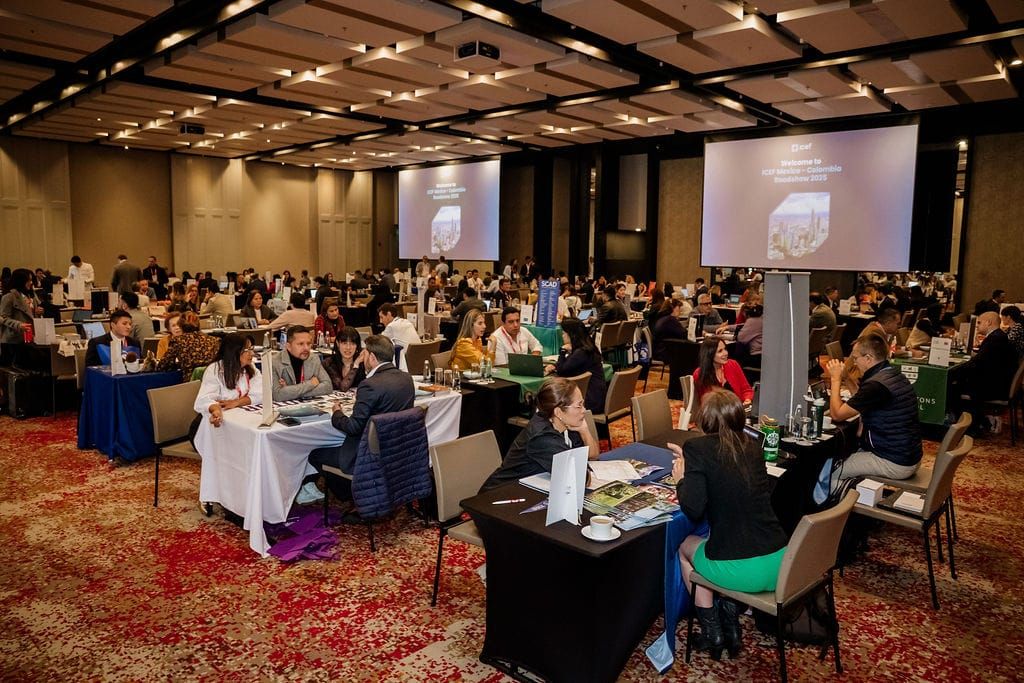 Professional conference attendees sit at round tables in a large ballroom with projector screens displaying presentations.