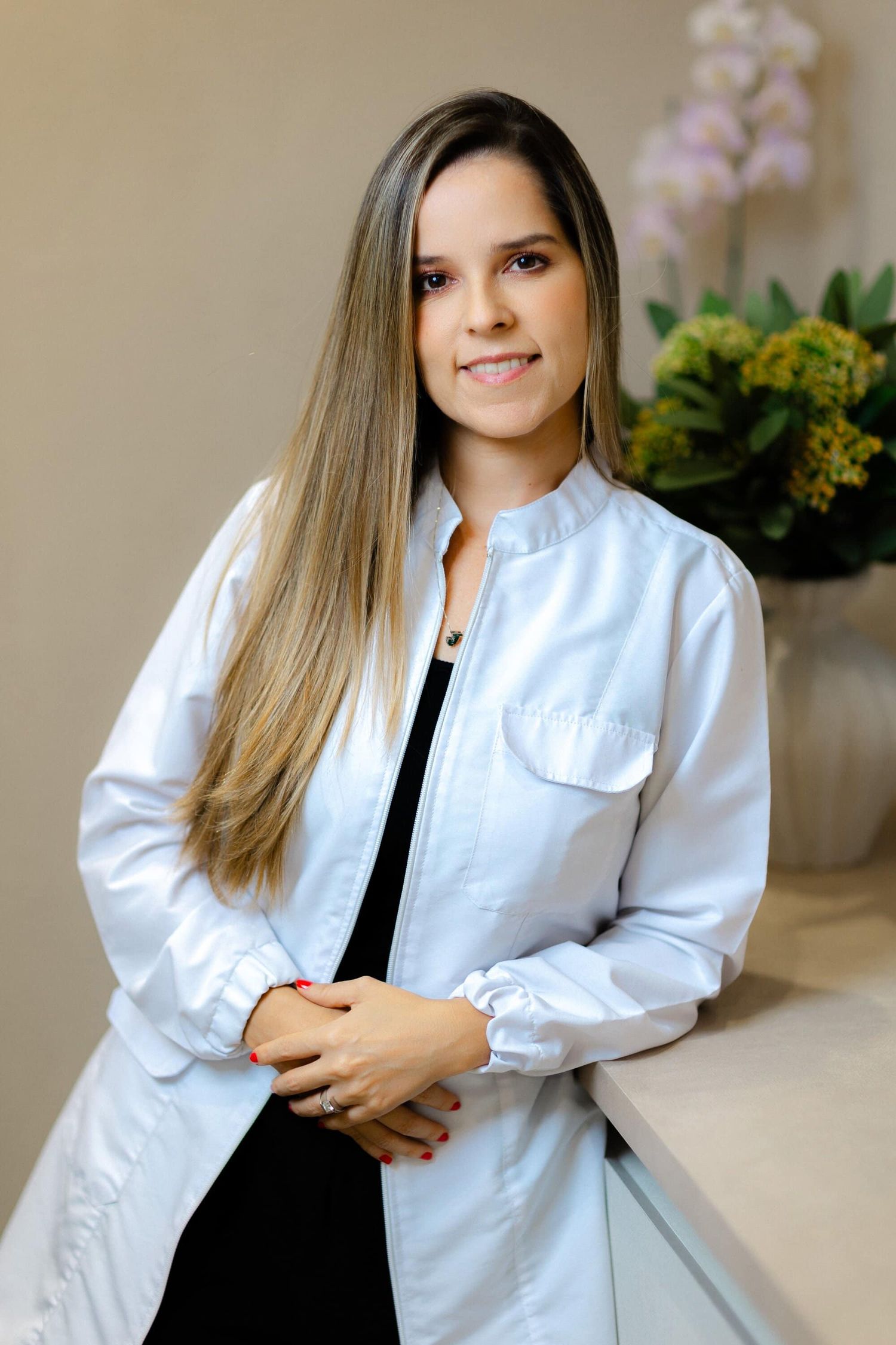 Professional wearing a crisp white medical coat over black attire stands in front of decorative plants and flowers.