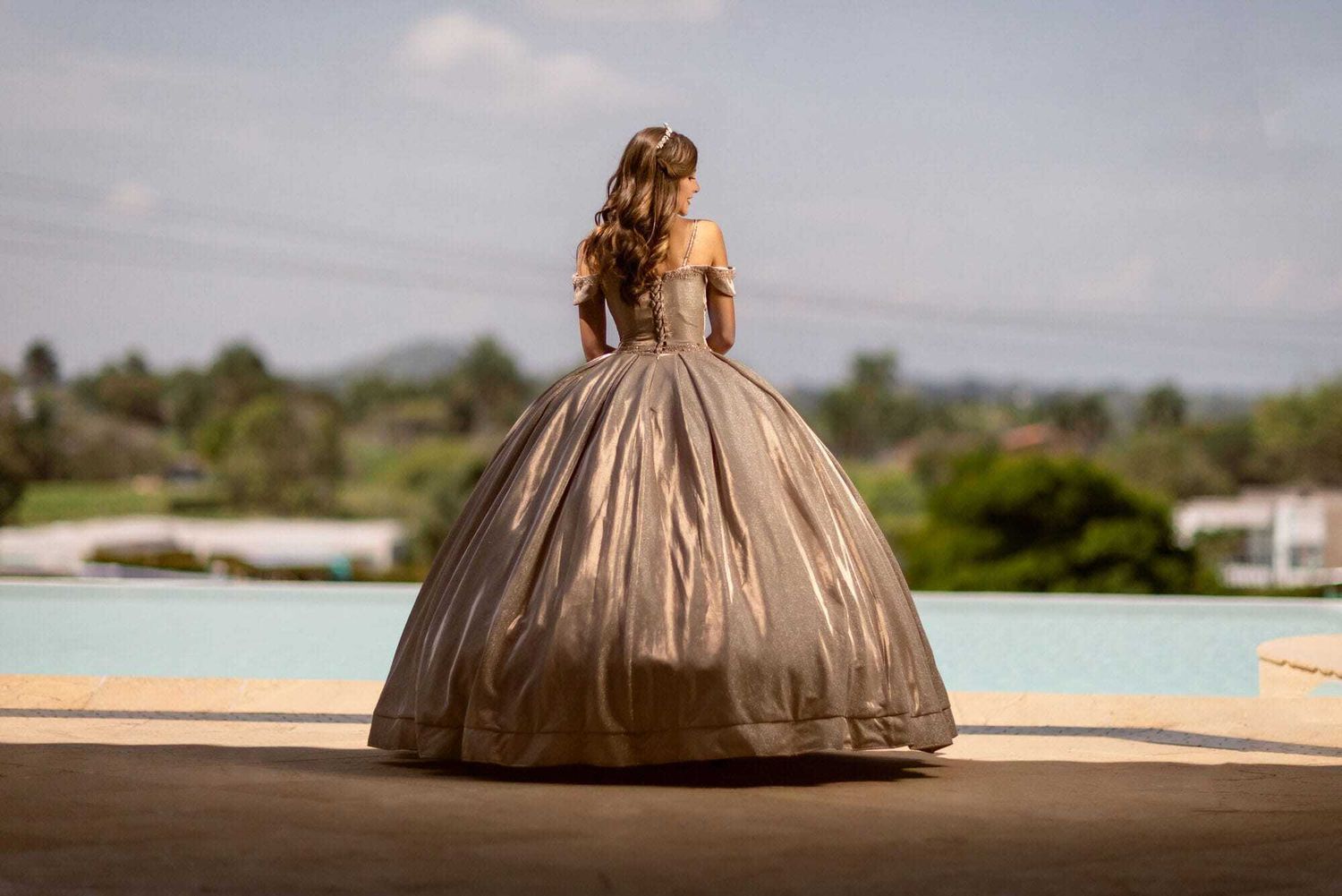 A figure in an elegant champagne-colored ball gown with a full skirt stands by a pool against a scenic landscape.