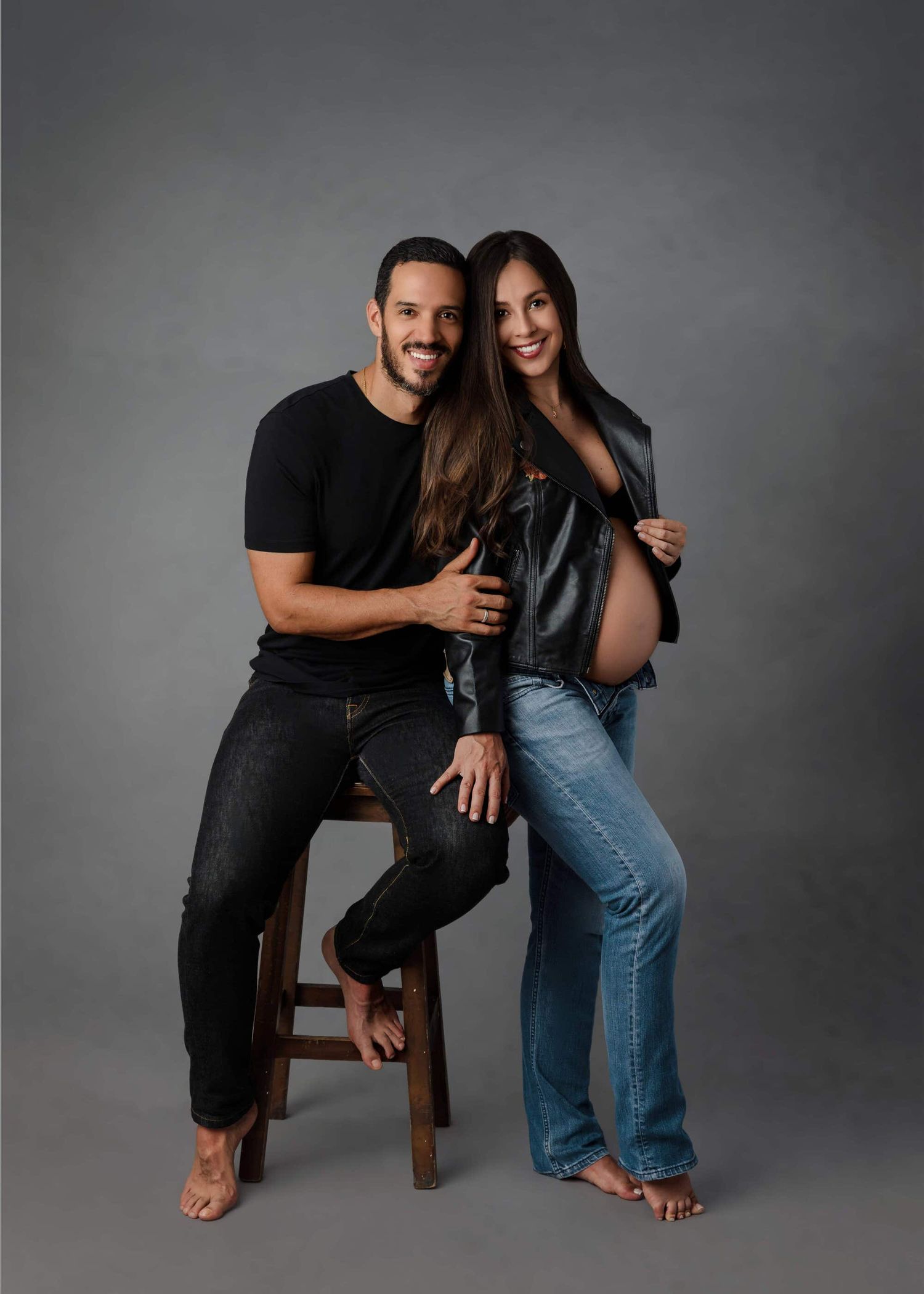 Professional maternity photo of couple wearing black and denim posing together on wooden stool against grey background.