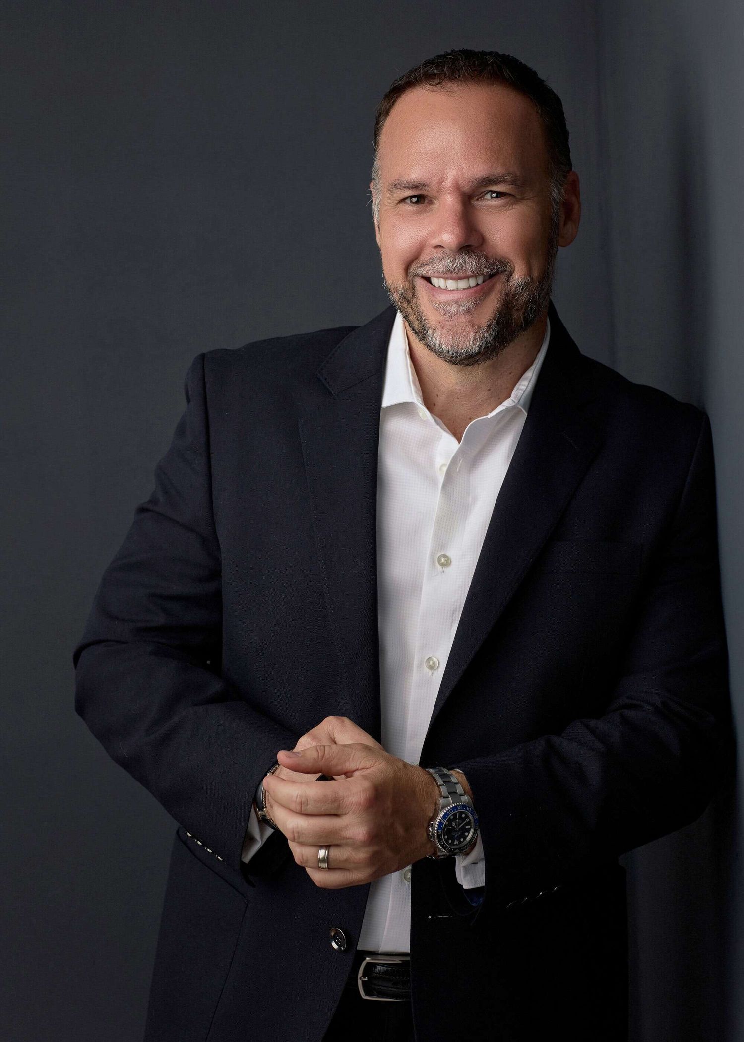 Professional business portrait of a person in a dark suit and white shirt against a gray background smiling warmly.