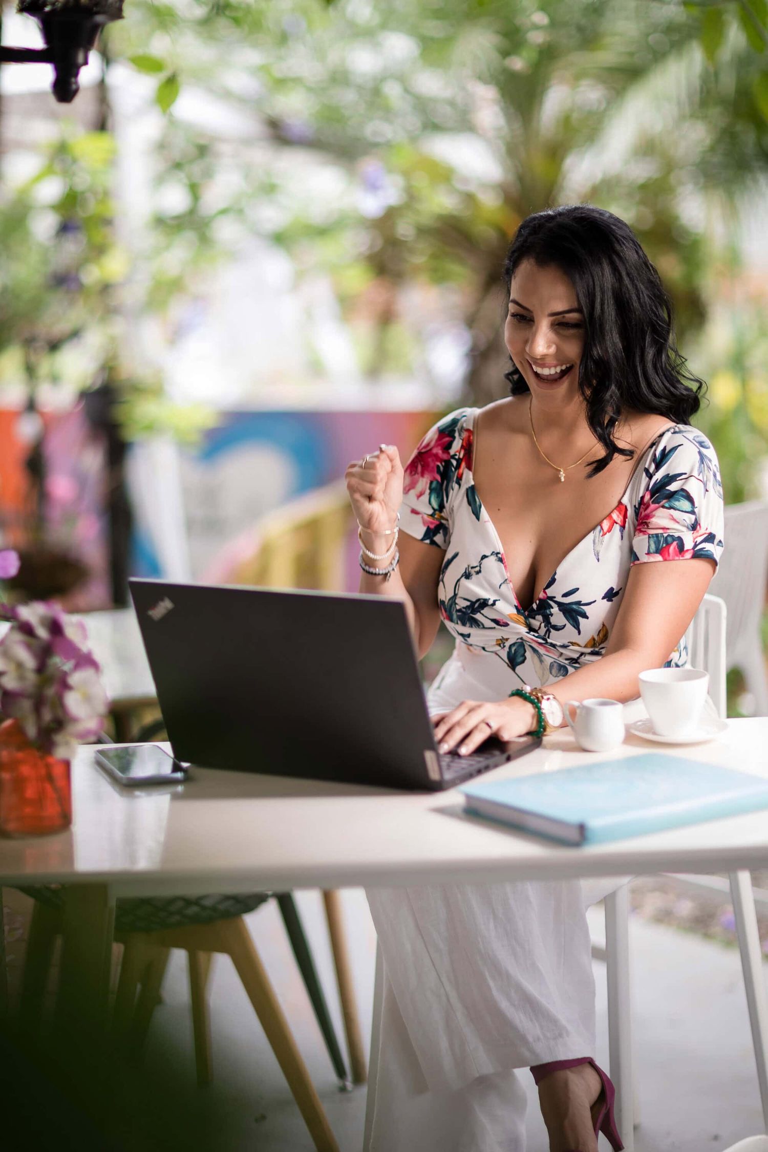 A woman in a floral dress works on a laptop at a bright cafe table with coffee and notebook.