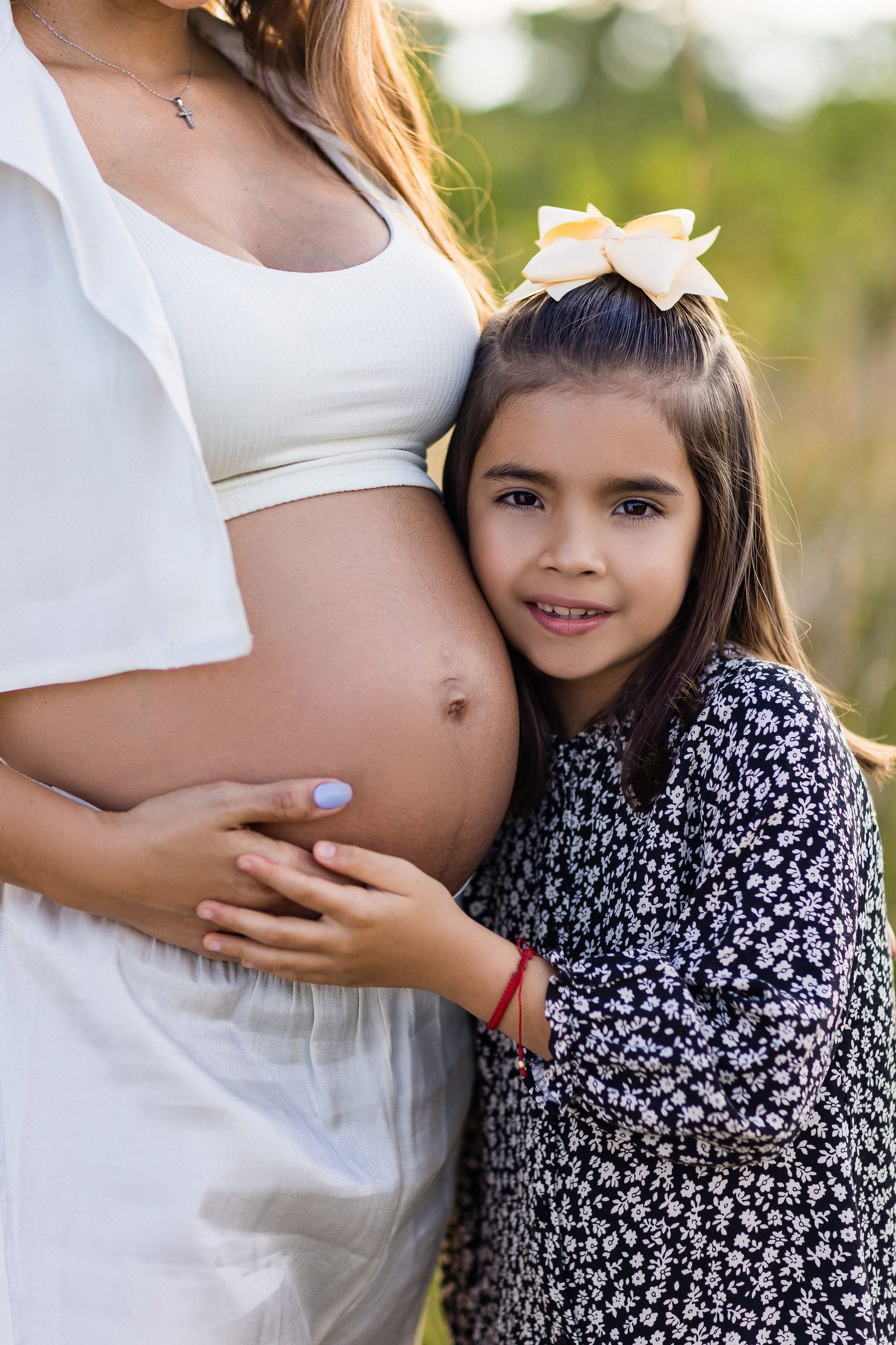 Young child in floral dress hugs a pregnant woman's belly while both stand outdoors in natural lighting.