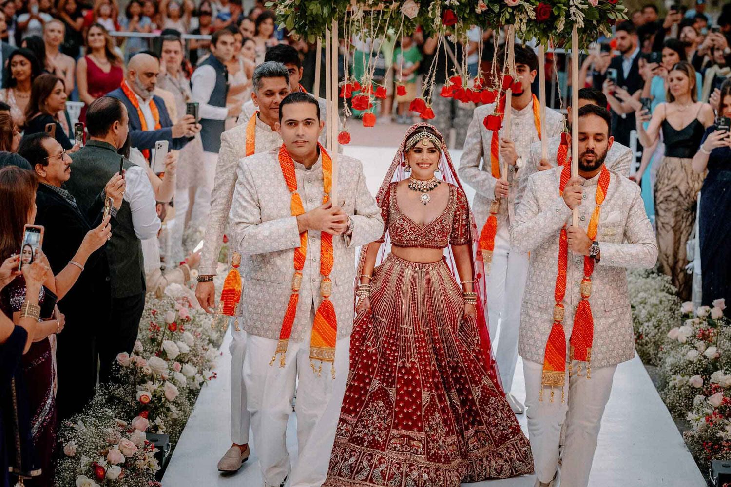 A bride in a traditional red lehenga walks down the aisle with groomsmen in white suits at an Indian wedding ceremony.