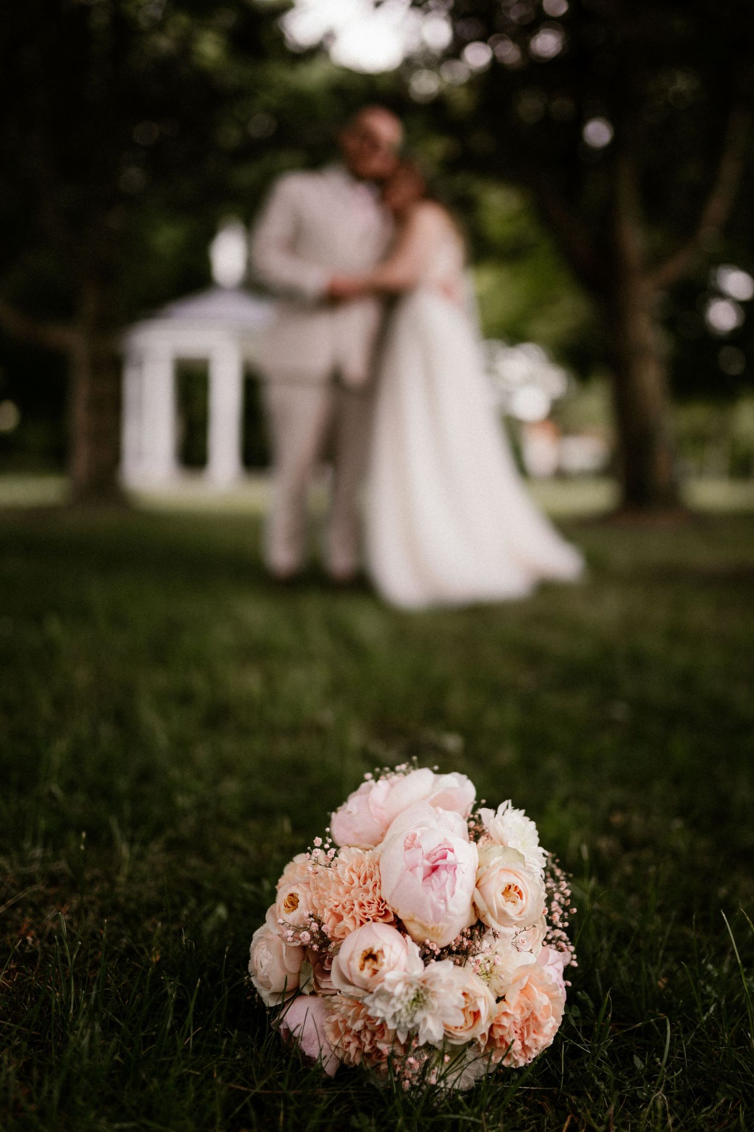 A delicate blush and cream peony bouquet lies on lush grass with soft bokeh in the background.
