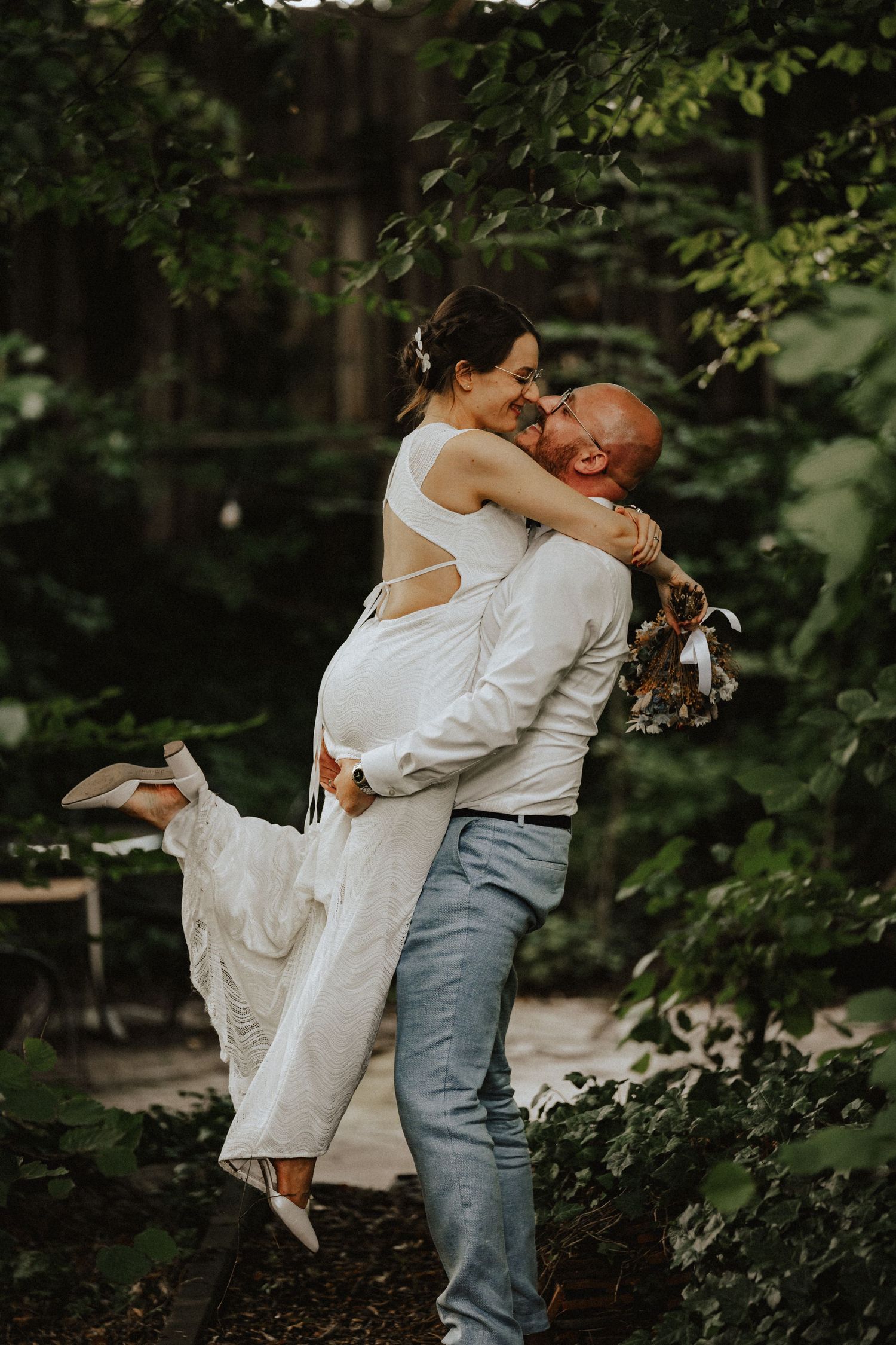 A romantic couple embraces in a forest setting with the bride wearing a flowing white dress and groom in casual attire.