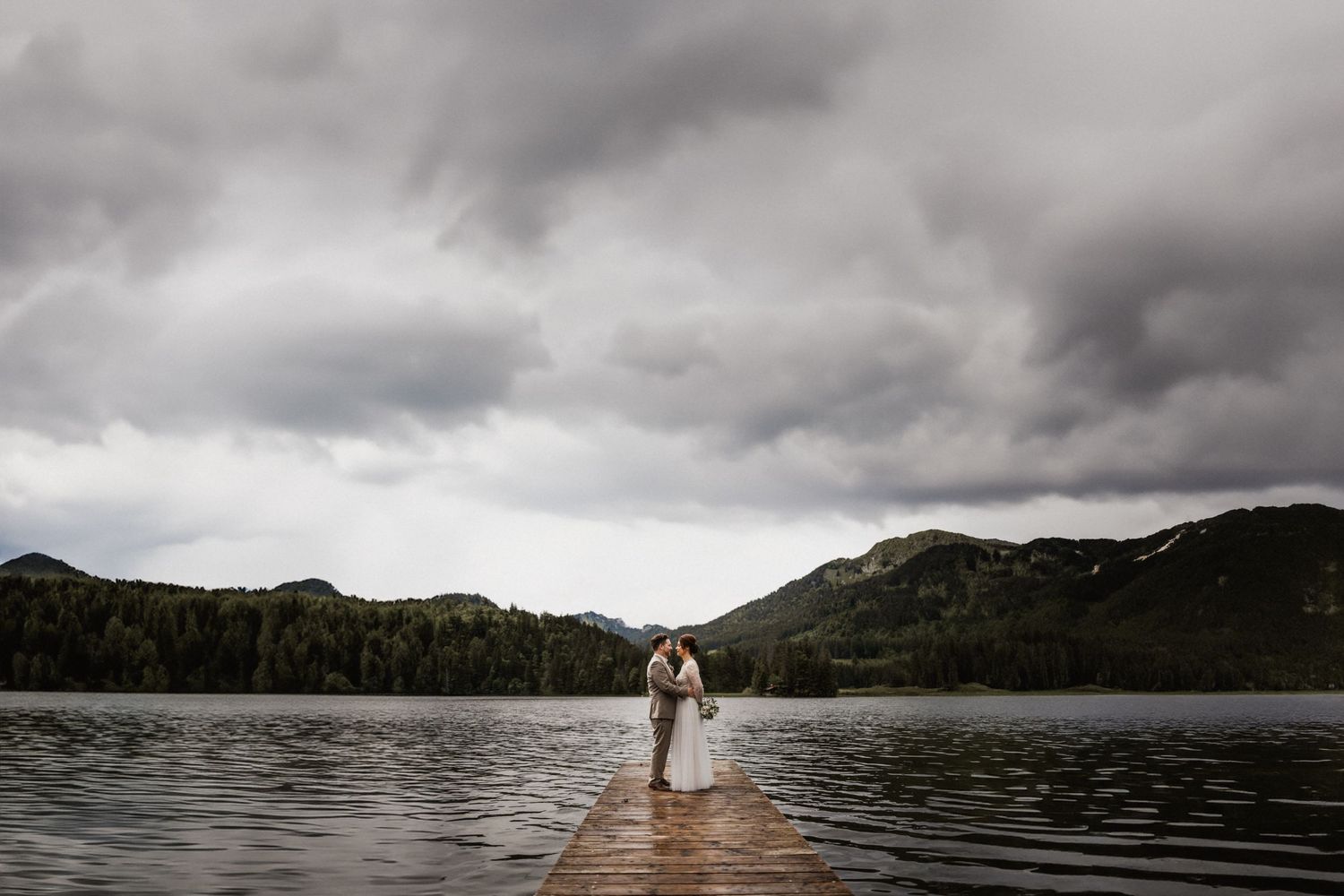 A couple stands at the end of a wooden dock on a mountain lake under dramatic storm clouds.