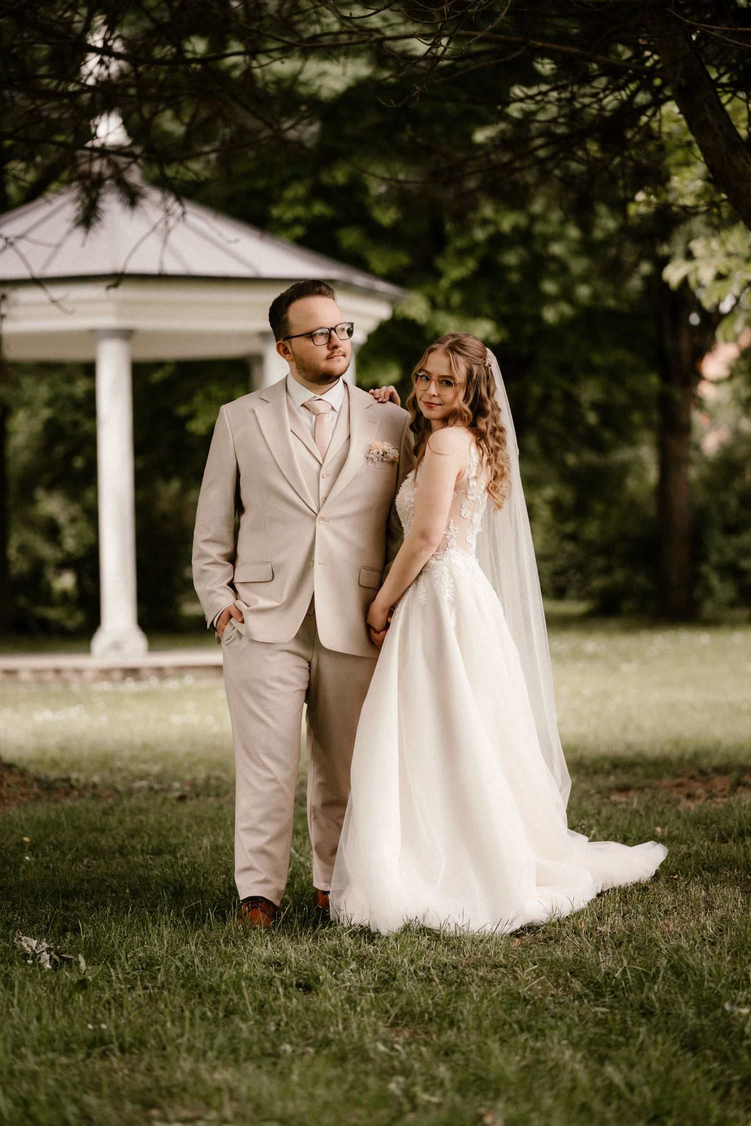 A couple in wedding attire poses together in front of a white gazebo on a lush green lawn.