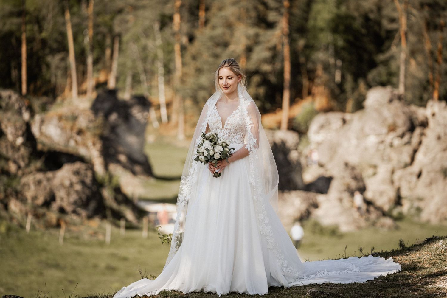 A bride in a flowing white lace gown and veil holds a bouquet while standing in a scenic wooded area with rocky outcrops.