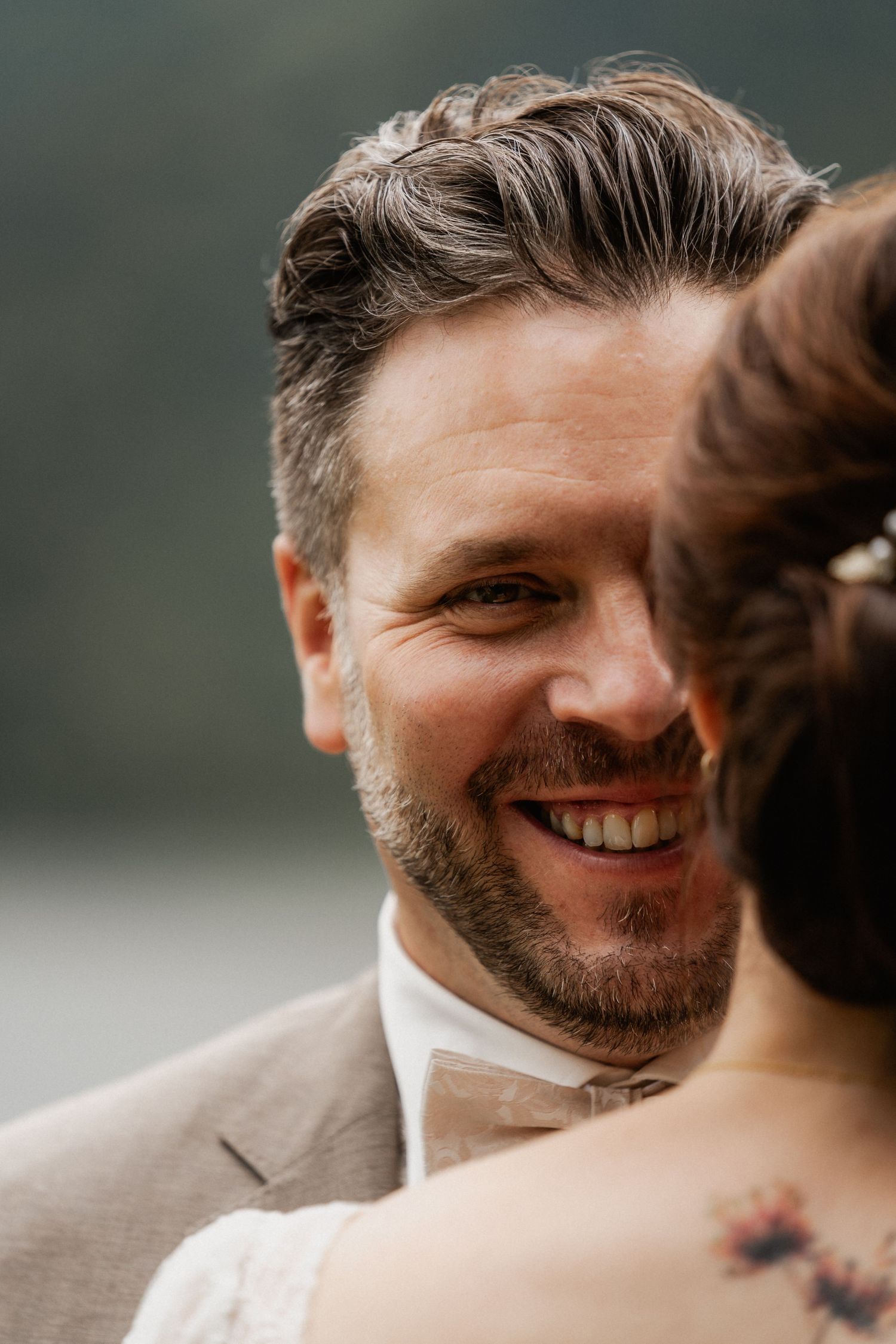 A person in a beige suit and bowtie smiles warmly during what appears to be a wedding moment.