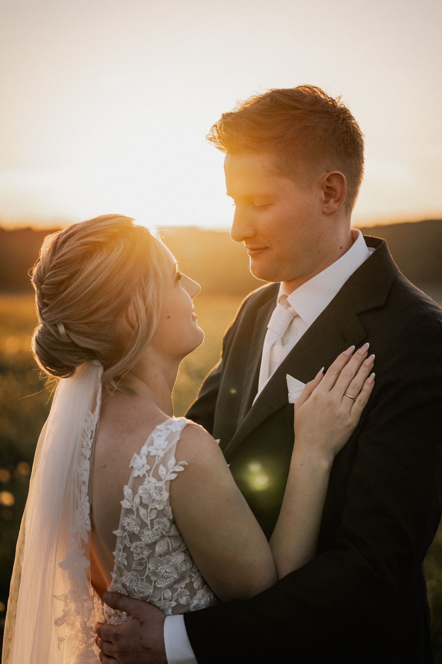 Romantic sunset wedding portrait with bride in lace dress and groom in black suit embracing during golden hour.