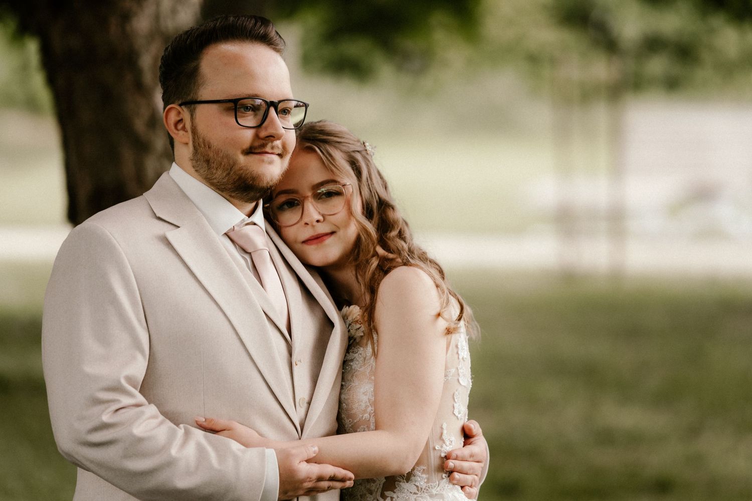 A romantic outdoor wedding portrait shows a couple embracing in soft natural lighting, with the groom in a beige suit.