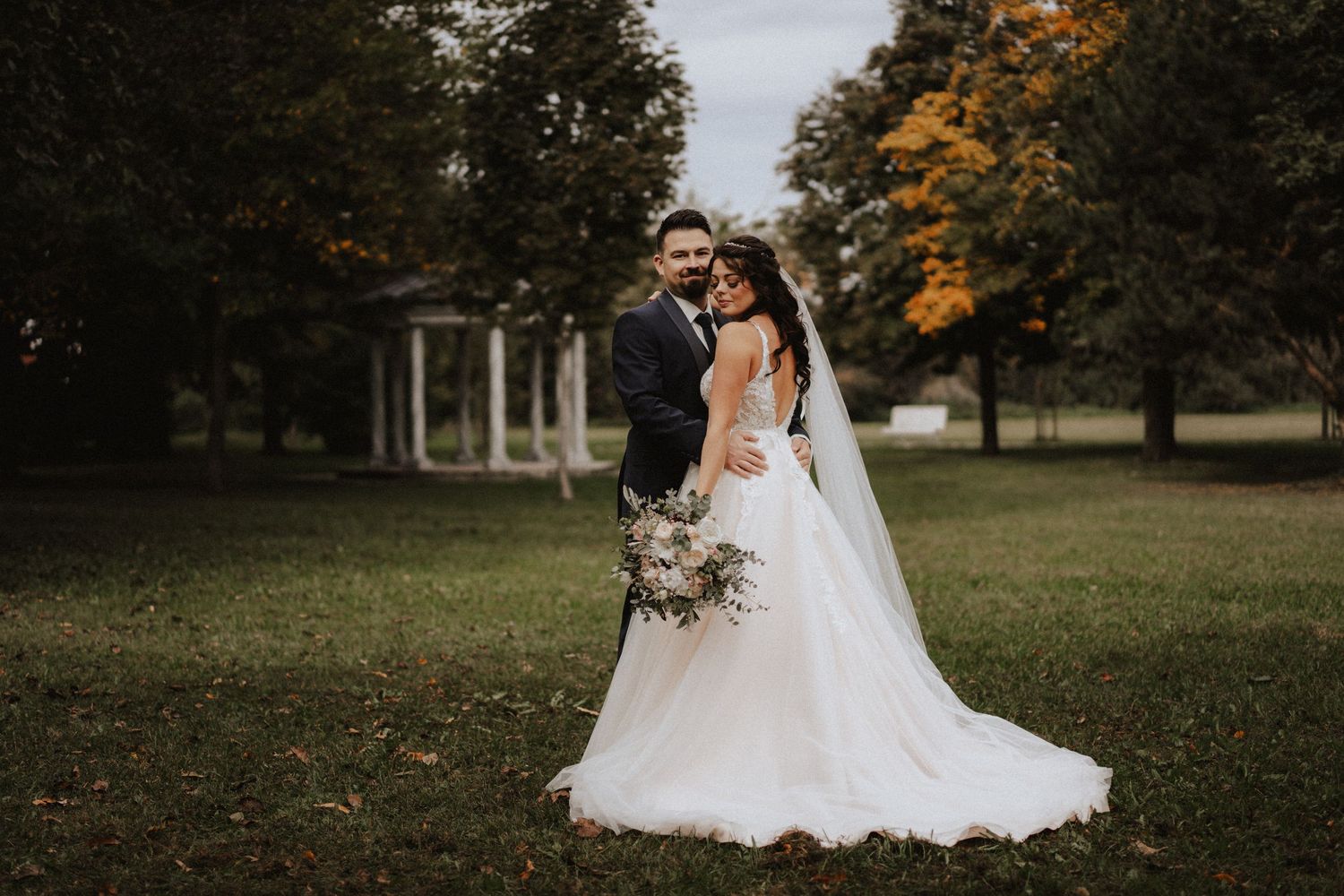 Newlyweds standing together on grass lawn with autumn trees and white gazebo in background at outdoor wedding venue.