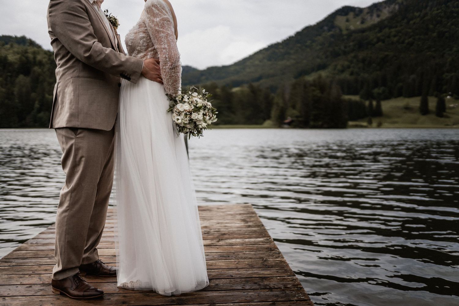 A couple in wedding attire stands on a wooden dock by a serene mountain lake, holding a white floral bouquet.