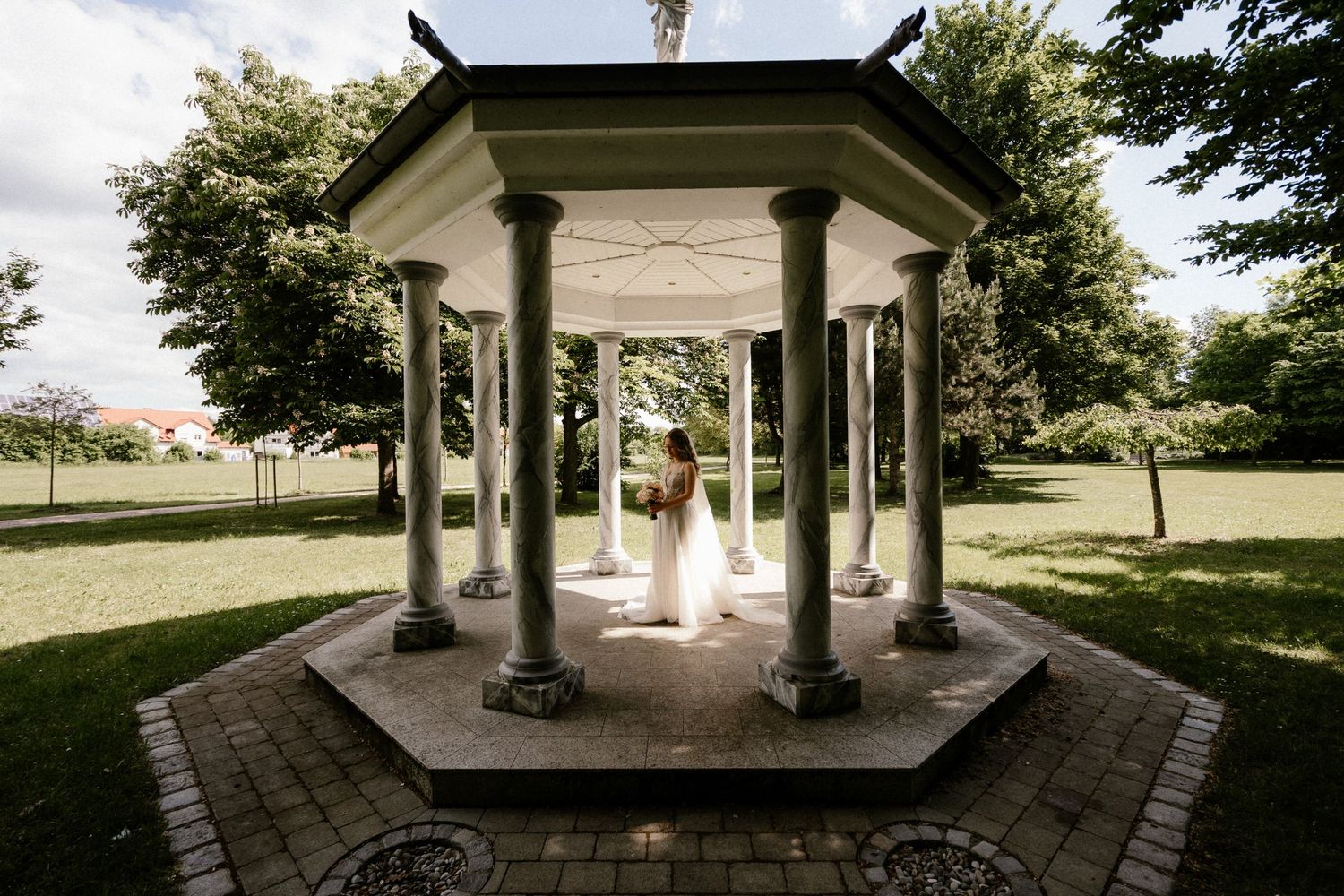 A vintage-style hexagonal gazebo with white pillars sits on a brick patio surrounded by green lawns and trees.