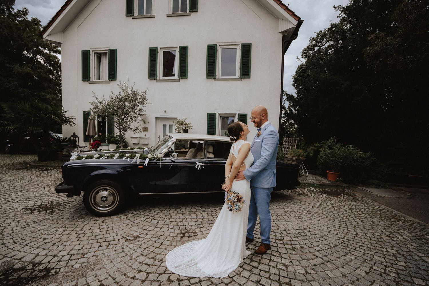Elegant couple stands by vintage black car on cobblestone driveway in front of white traditional house.