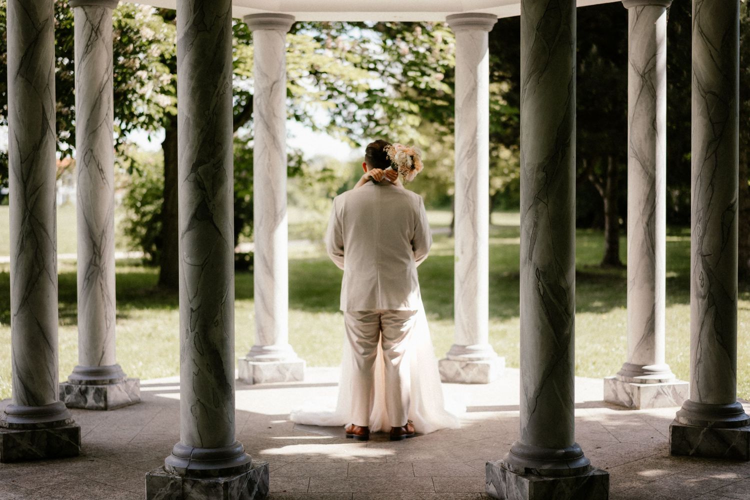 A person wearing beige clothing stands between elegant stone columns overlooking a garden setting.