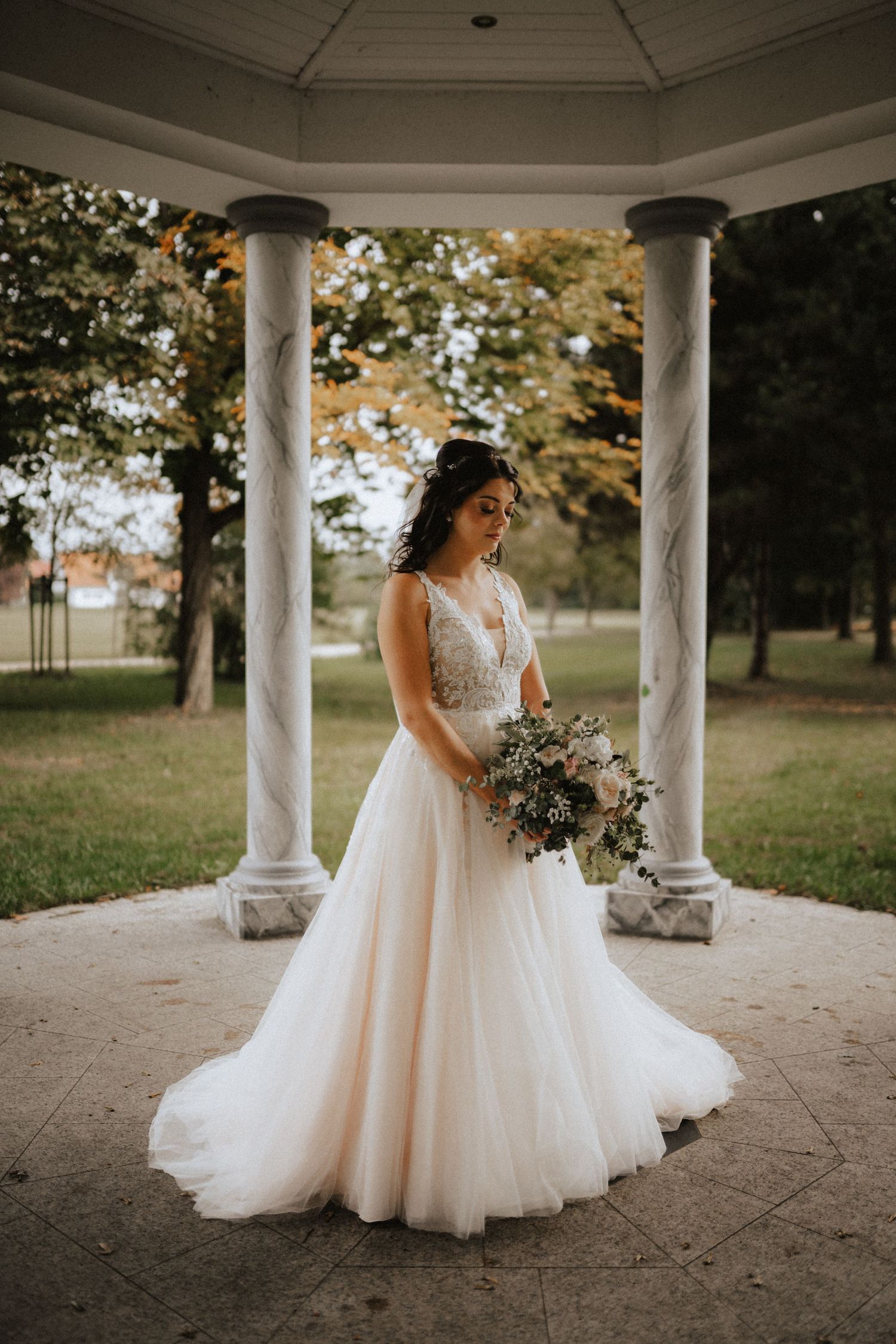 A bride in an elegant lace and tulle wedding gown stands between white columns holding a rustic floral bouquet.