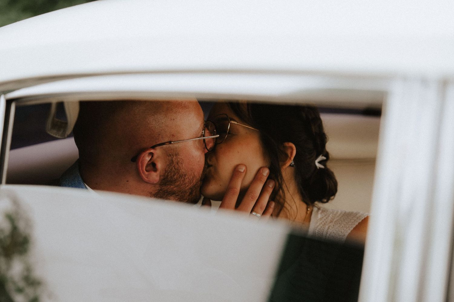 A romantic couple shares an intimate kiss viewed through a car window frame, creating a tender candid moment.