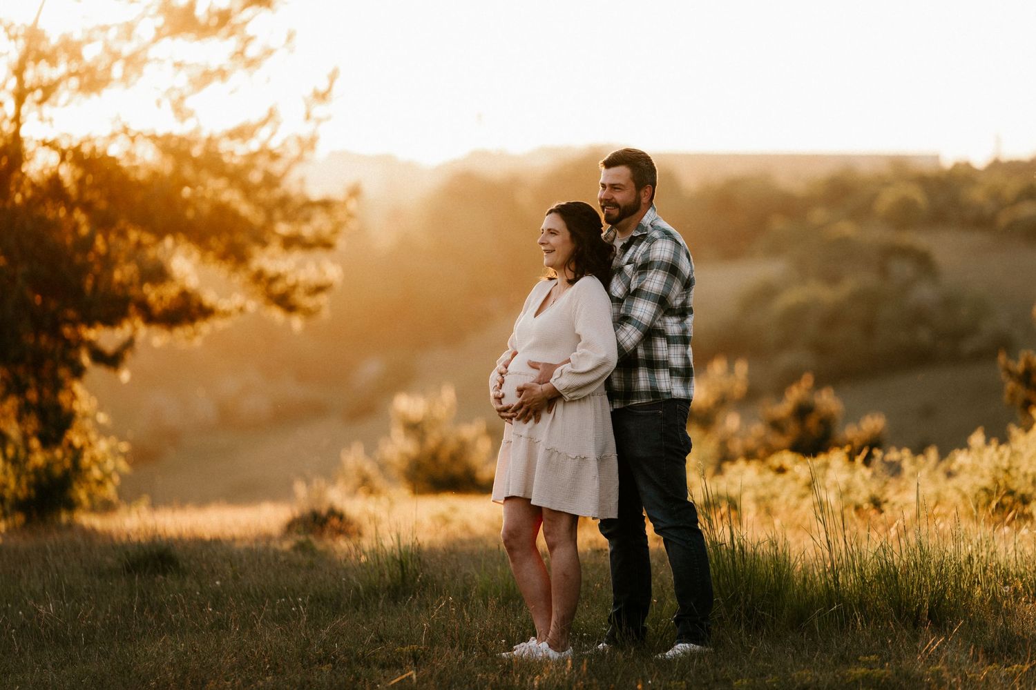 A couple embraces while standing in a golden sunlit meadow at sunset during a romantic outdoor photoshoot.