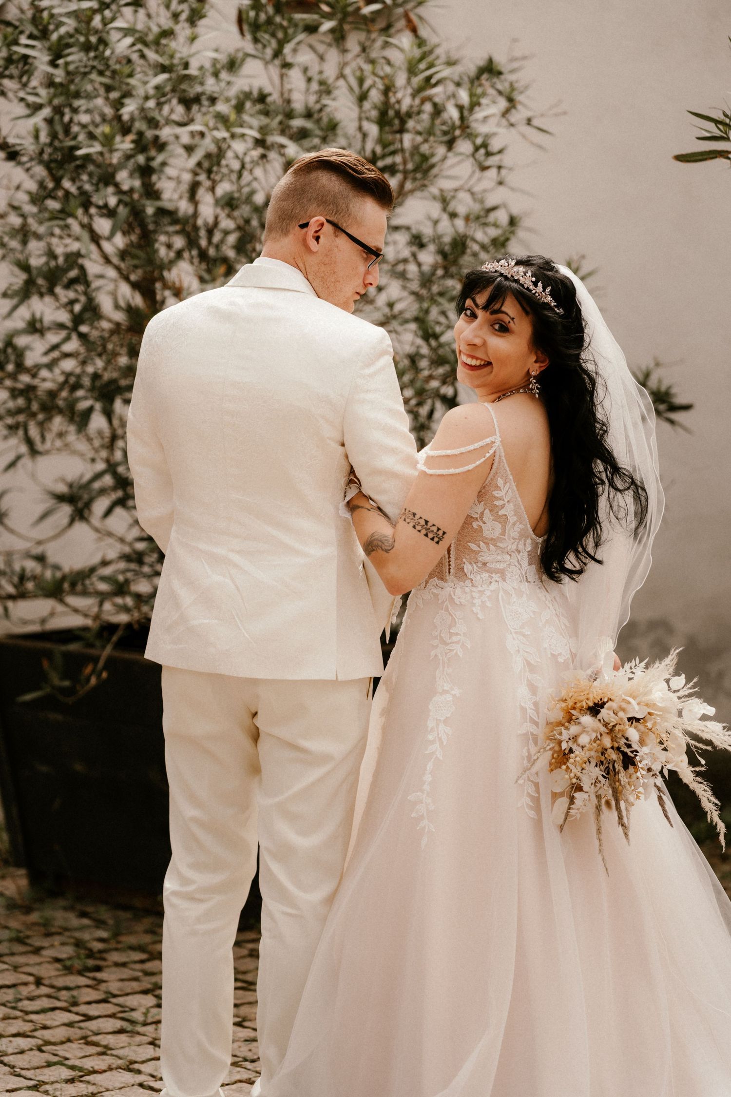 A couple in white wedding attire share a candid moment outdoors while the bride holds a neutral-toned dried floral bouquet.