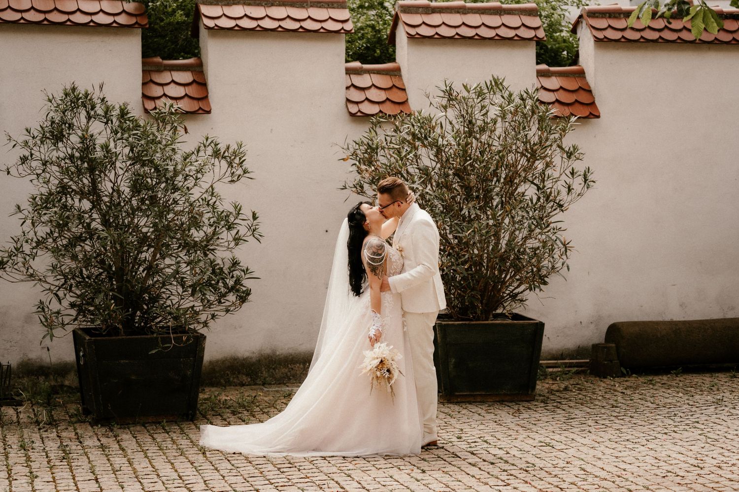 Two people share an intimate moment in their wedding attire beside potted olive trees against a stucco wall.