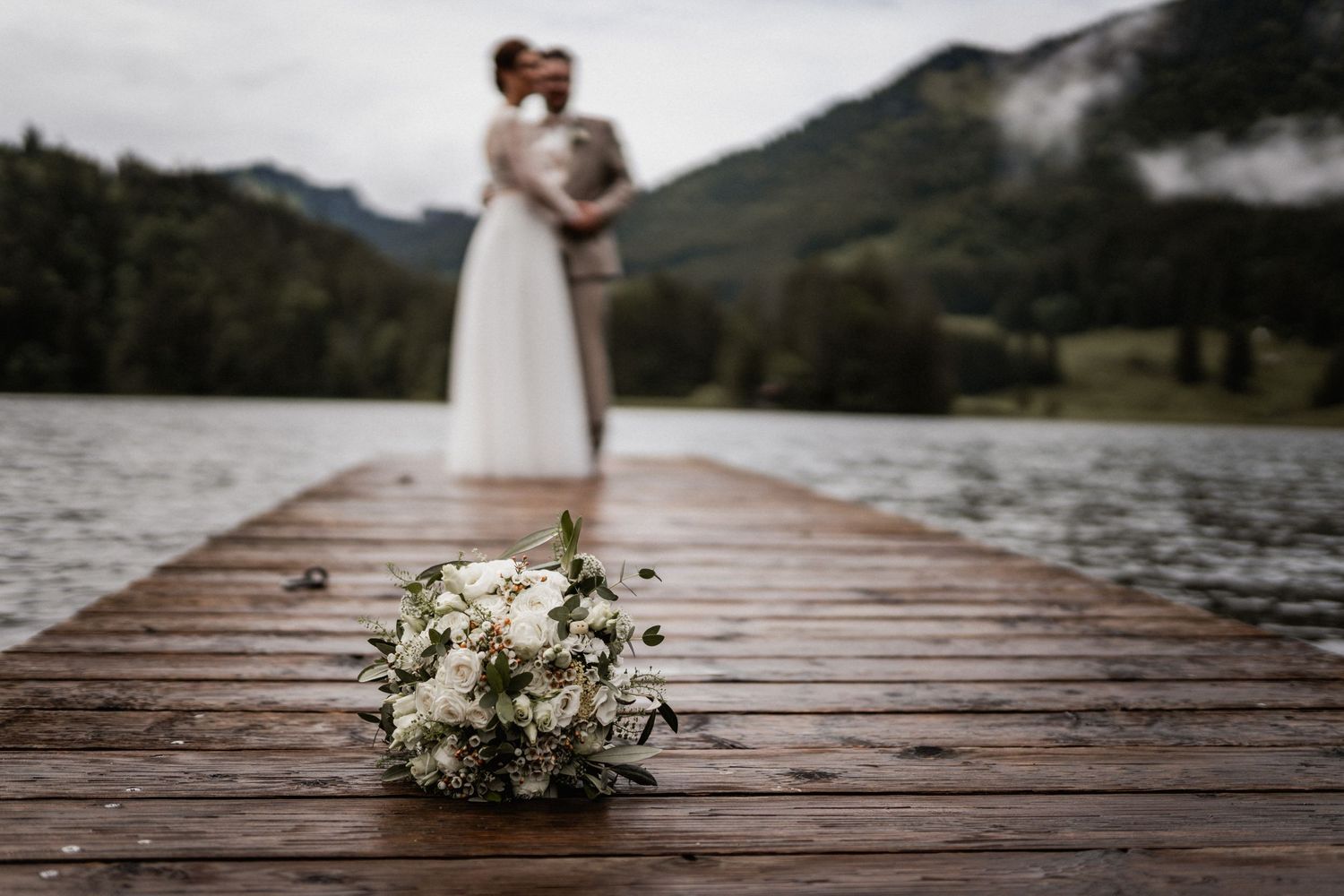 A white wedding bouquet lies on a wooden dock overlooking a misty mountain lake with a couple embracing in soft focus.