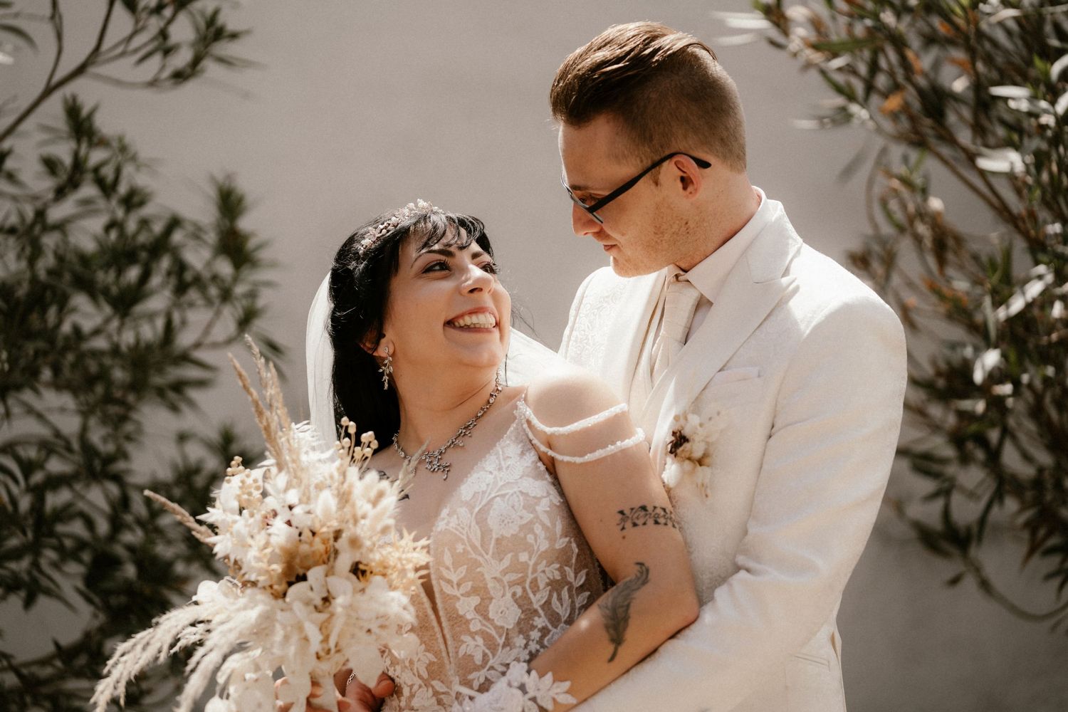 Happy couple in white wedding attire share a joyful moment while holding a dried floral bouquet against white wall.