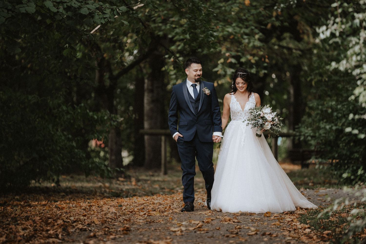 Couple in wedding attire walks through a scenic autumn forest setting while holding hands during their photoshoot.