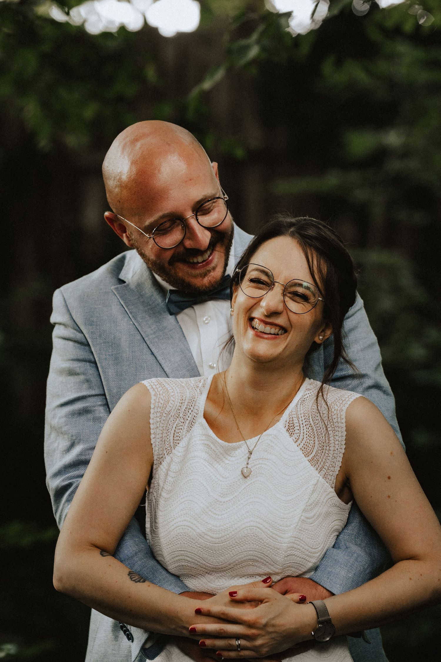 A romantic outdoor wedding photo showing a couple wearing glasses embracing and laughing together in a natural setting.