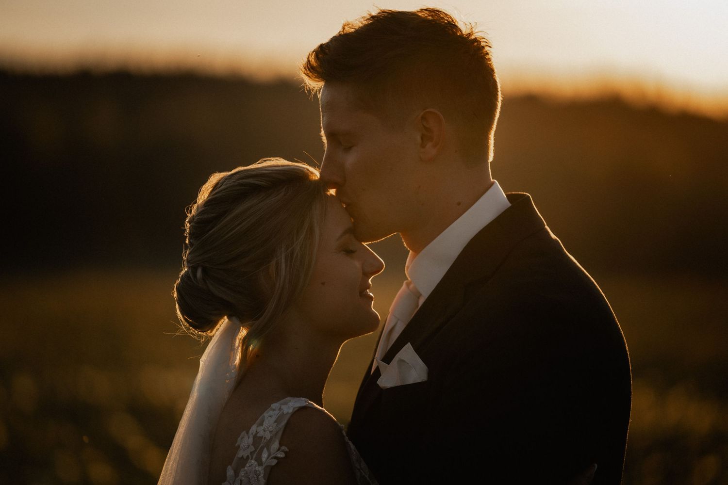 A romantic wedding couple embraces in warm evening sunlight against a rural landscape.