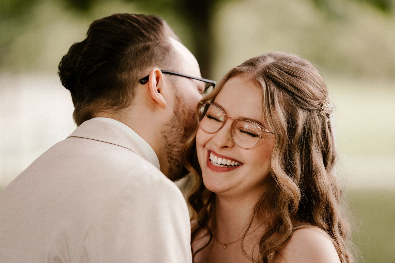 A couple wearing glasses share an intimate moment outdoors, both smiling joyfully with wavy brown hair and light attire.