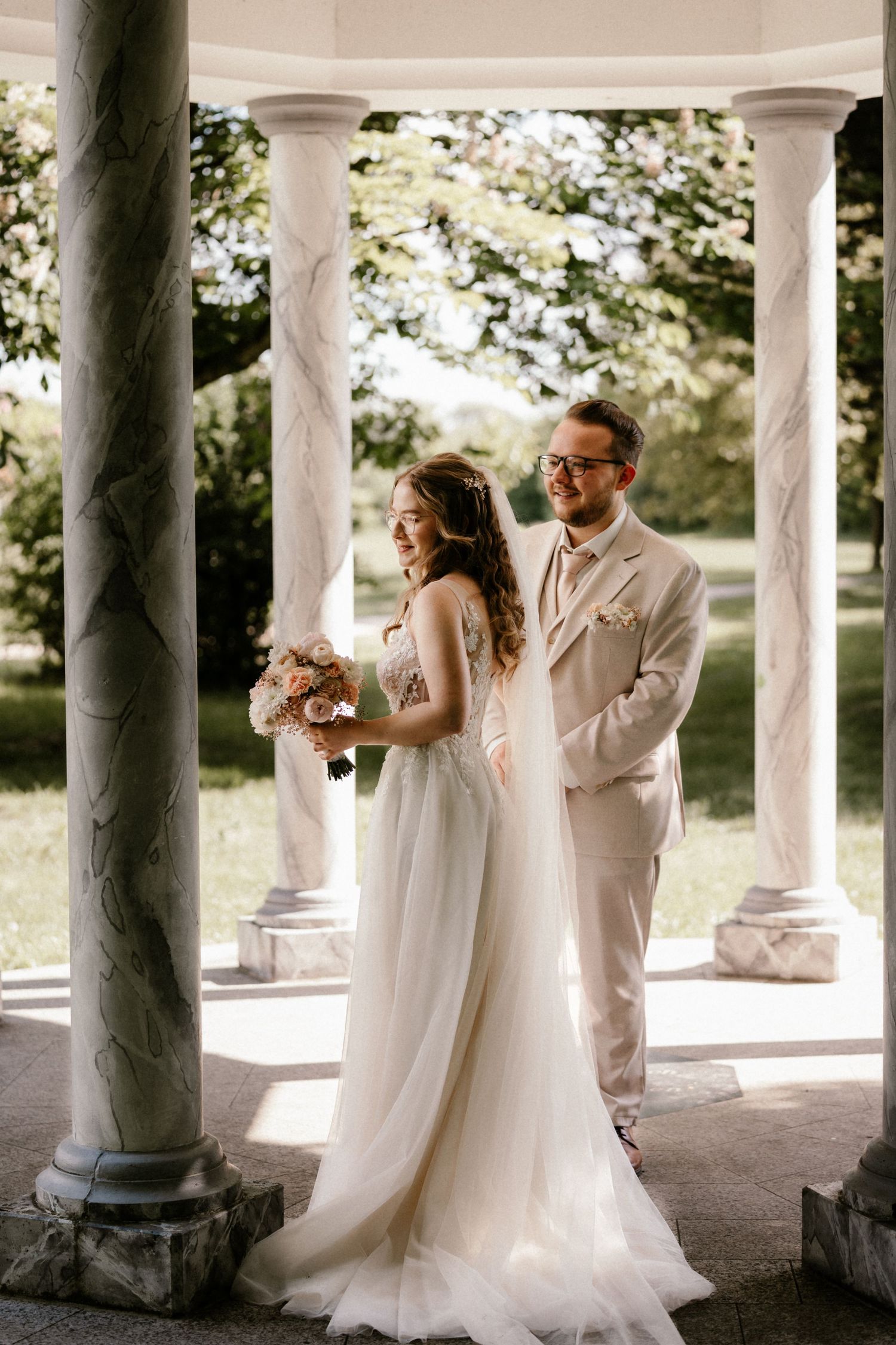 Newlywed couple poses romantically under classic white stone gazebo with columns in soft natural lighting.