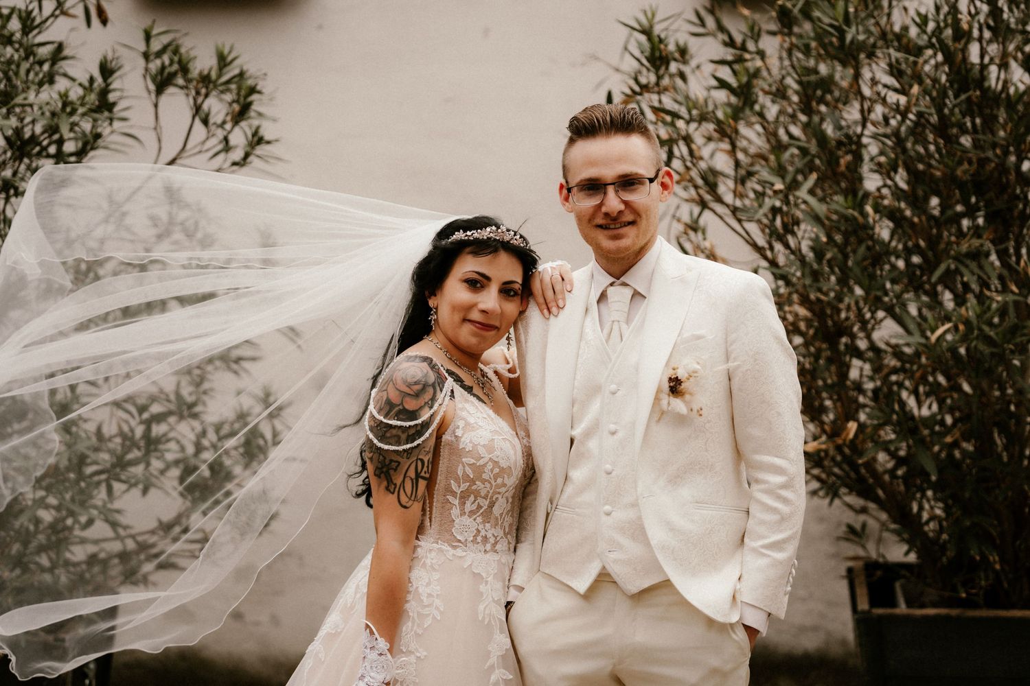 A wedding couple poses outdoors with a flowing veil against a garden backdrop.